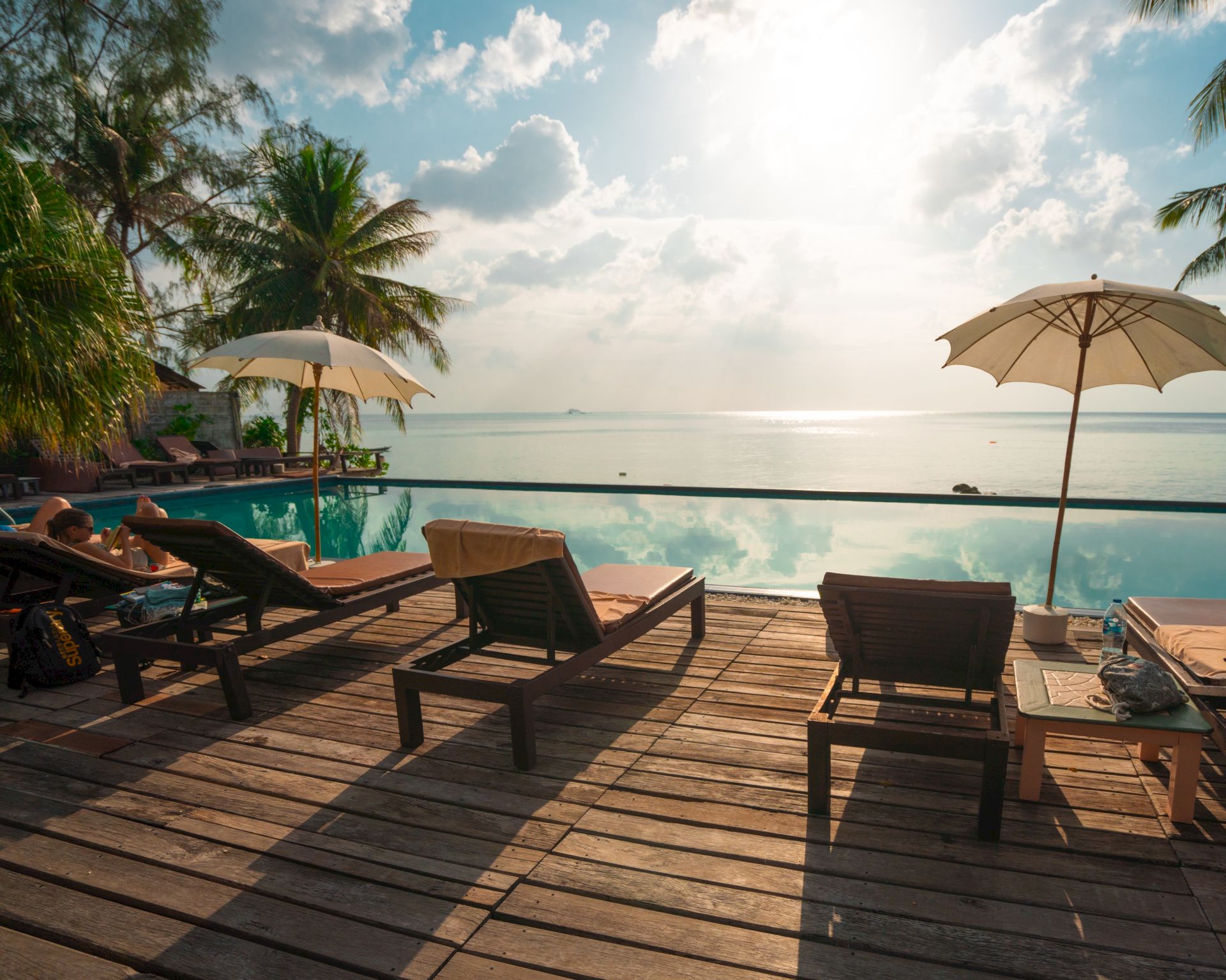 A serene tropical setting with sun loungers and umbrellas by a pool overlooking the ocean, with palm trees and a bright sky in the background.