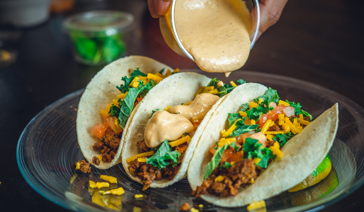 Three tacos on a plate being topped with creamy sauce. They are filled with ground meat, cheese, lettuce, and diced tomatoes.