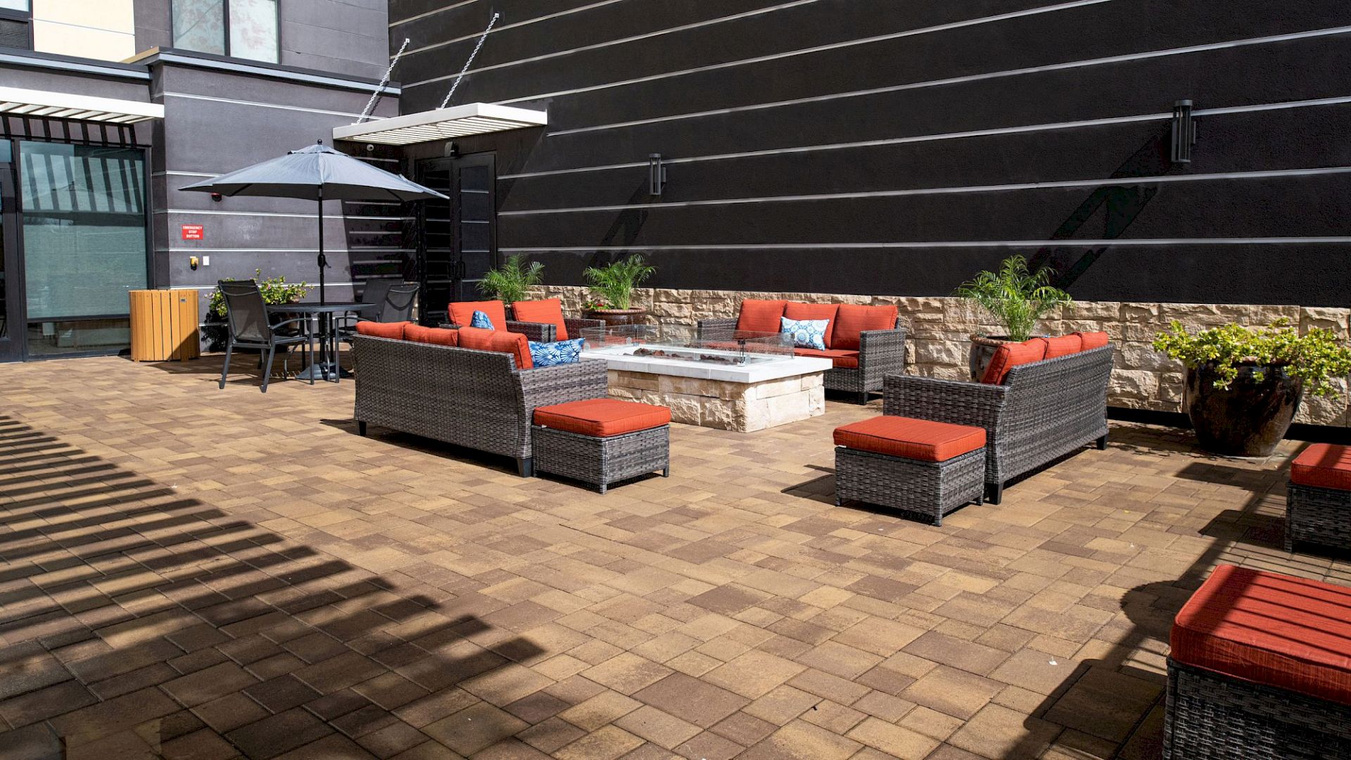 An outdoor patio with cushioned wicker sofas, a fire pit, potted plants, and a table with chairs under an umbrella, on a brick floor.