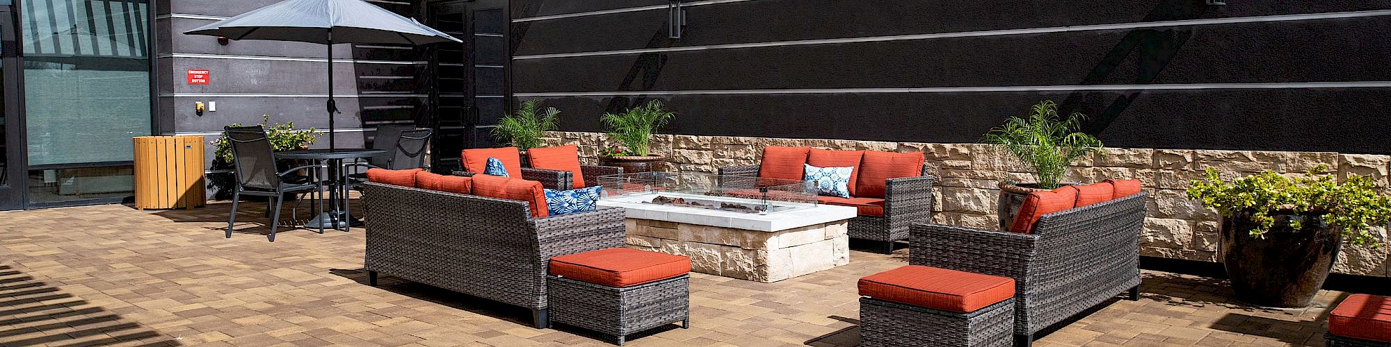 An outdoor patio with cushioned wicker sofas, a fire pit, potted plants, and a table with chairs under an umbrella, on a brick floor.