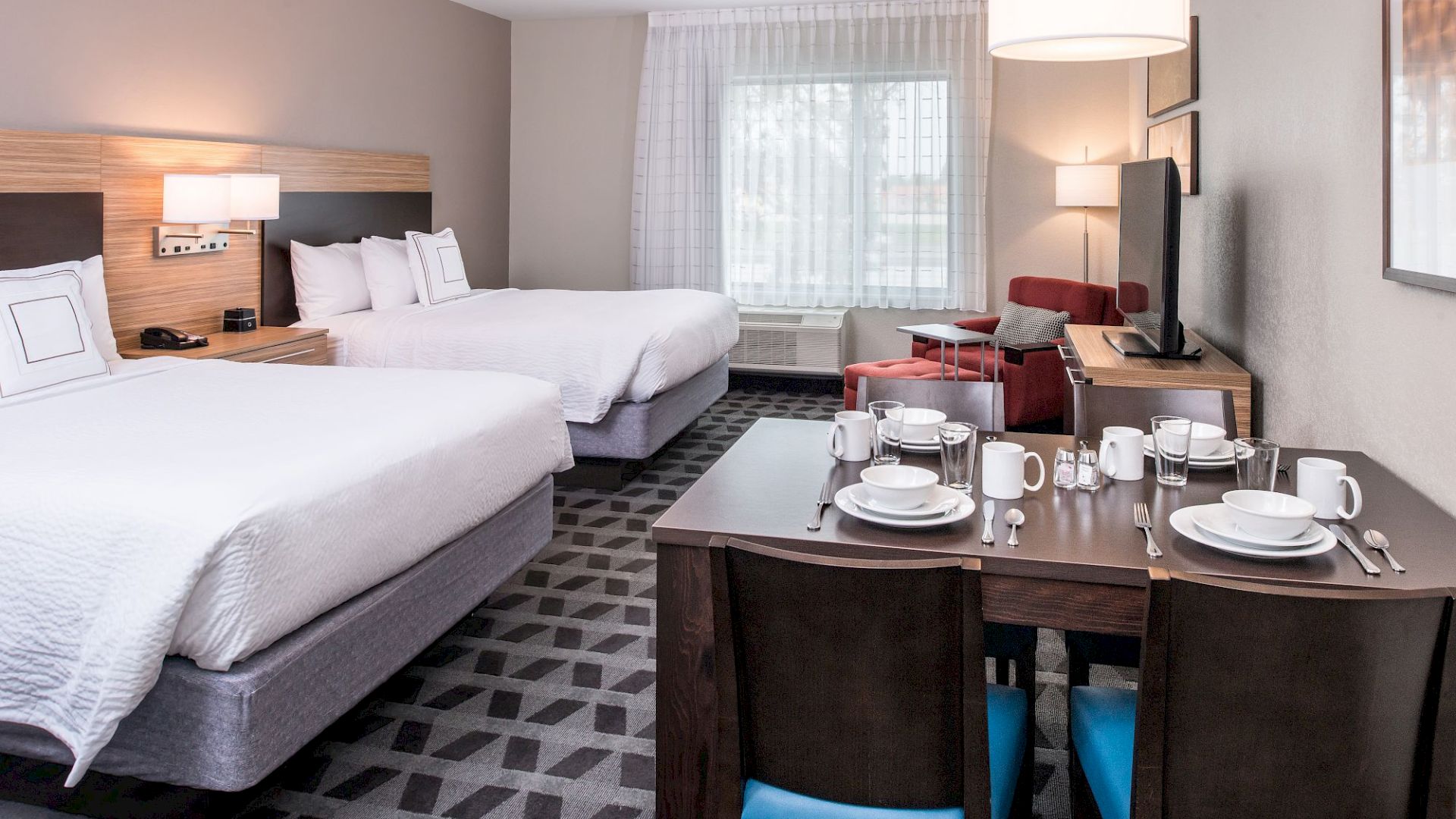 A hotel room with two beds, a dining table set for four, a TV, a red armchair, and large window with curtains. The overall decoration is modern.
