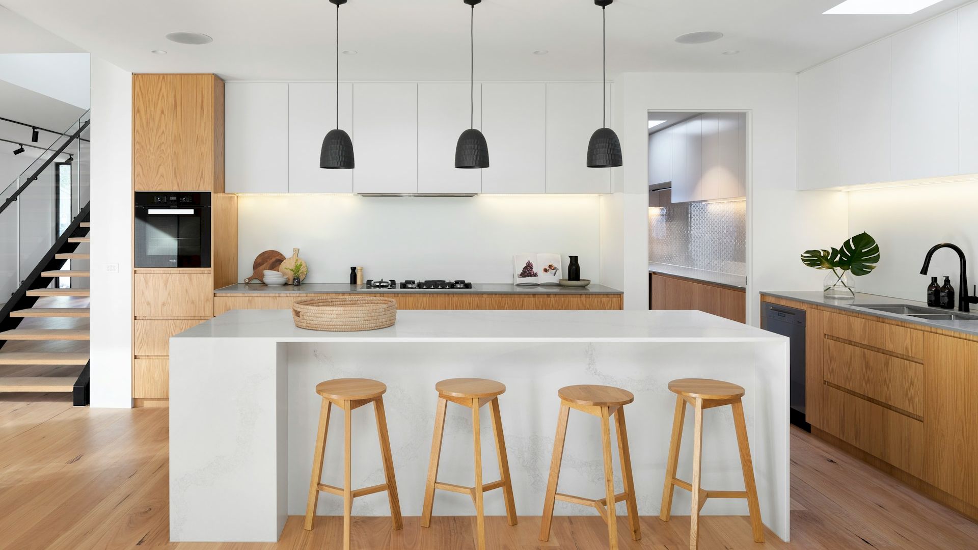A modern kitchen with white cabinets, wooden accents, a large island with stools, black pendant lights, a stainless steel sink, and an oven.