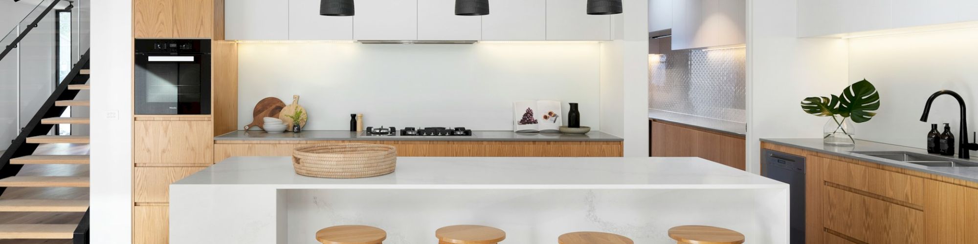 A modern kitchen with white cabinets, wooden accents, a large island with stools, black pendant lights, a stainless steel sink, and an oven.