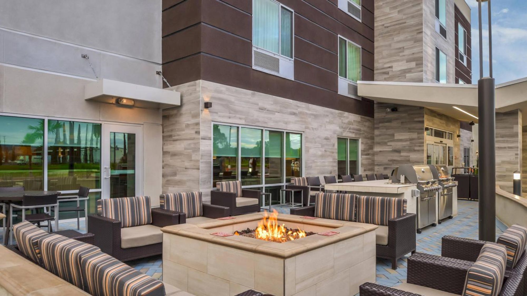 Outdoor patio with modern seating, a central fire pit, a grill area, and contemporary building facade in the background, ideal for social gatherings.