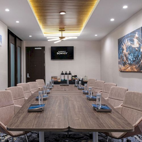 A modern conference room with a large table, cushioned chairs, water glasses, notebooks, and a screen displaying text. There is a coffee station at the back.