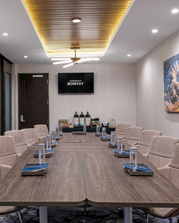 A modern conference room with a large table, cushioned chairs, water glasses, notebooks, and a screen displaying text. There is a coffee station at the back.