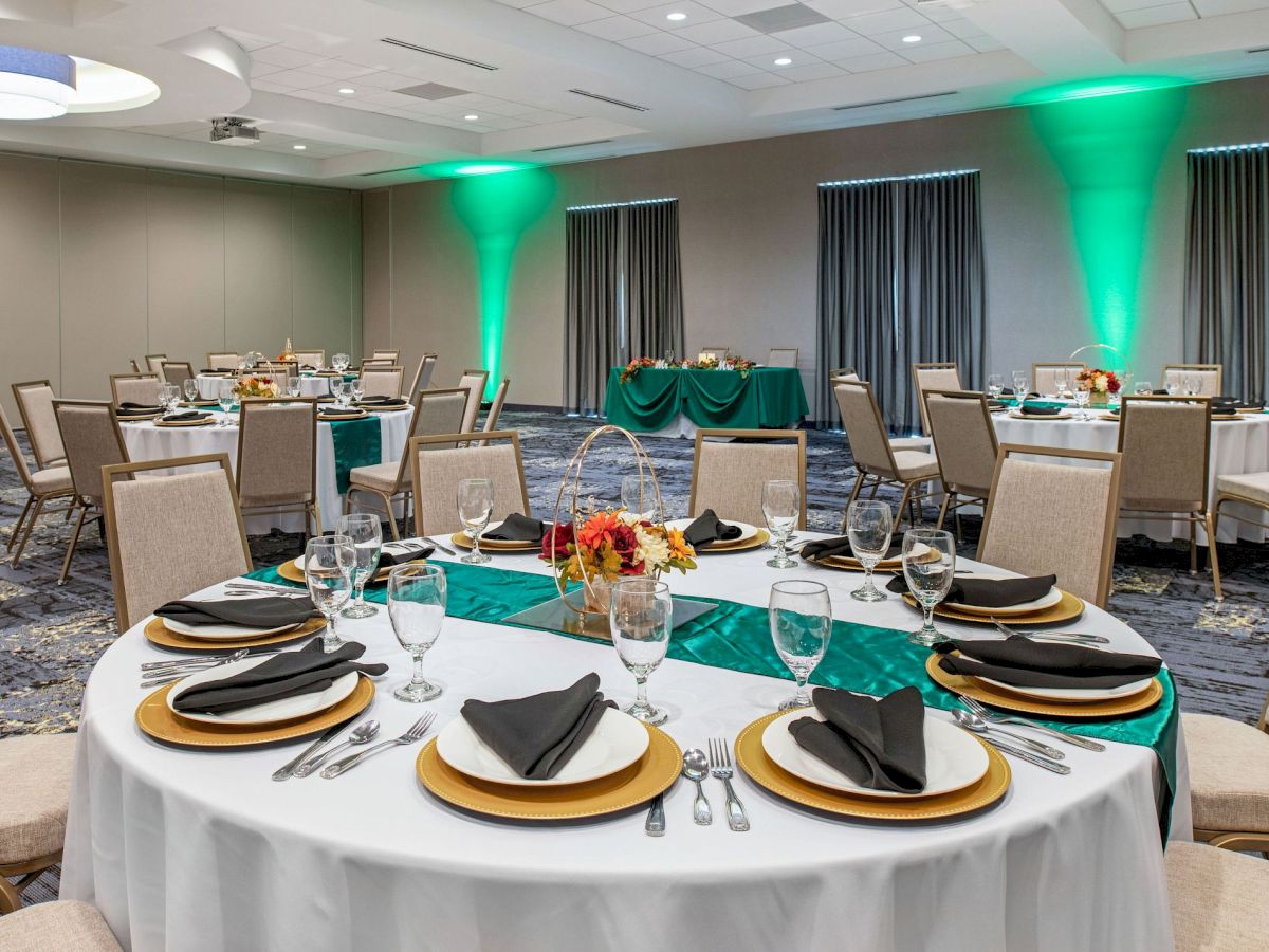 This image shows a banquet hall set up for an event with round tables, neatly arranged cutlery, black napkins on plates, and green lighting.