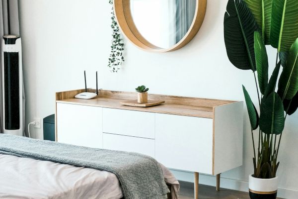 A modern bedroom with a bed, a wooden dresser with a circular mirror above it, and a large potted plant on the floor next to the dresser.