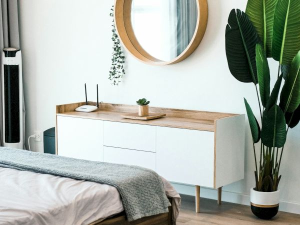 A modern bedroom with a bed, a wooden dresser with a circular mirror above it, and a large potted plant on the floor next to the dresser.