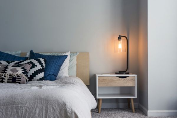 A cozy bedroom features a bed with blue and patterned pillows, a white duvet, and a bedside table with a lit modern lamp.