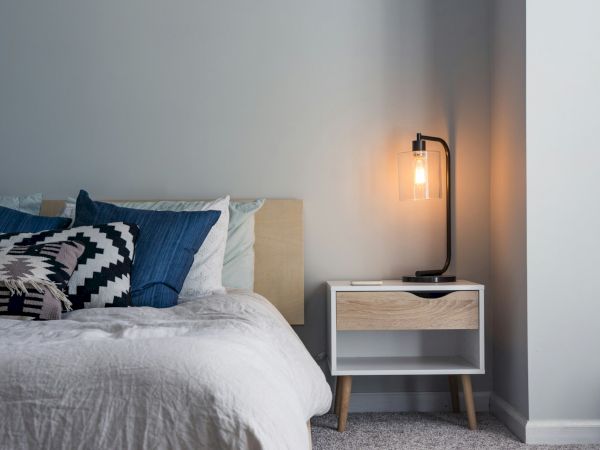 A cozy bedroom features a bed with blue and patterned pillows, a white duvet, and a bedside table with a lit modern lamp.