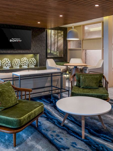 This image shows a modern living room with green armchairs, a round white coffee table, and a large television mounted on a stone wall.