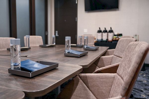 A conference room with a wooden table, chairs, water bottles, notebooks, and a TV on the wall. Beverage dispensers are in the background.