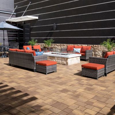 An outdoor patio seating area with a fire pit, wicker furniture with red cushions, an umbrella-covered table, and potted plants against a dark wall.