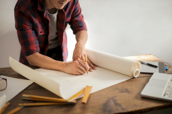 A person is reviewing and marking up large sheets of paper on a wooden desk, surrounded by pencils, a ruler, and a laptop, possibly working on a design.