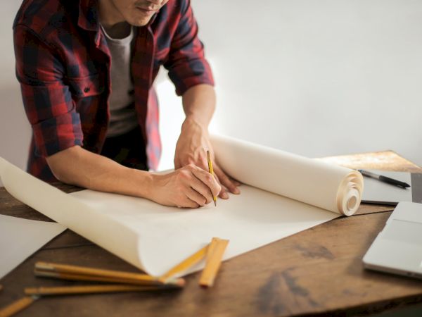 A person is reviewing and marking up large sheets of paper on a wooden desk, surrounded by pencils, a ruler, and a laptop, possibly working on a design.