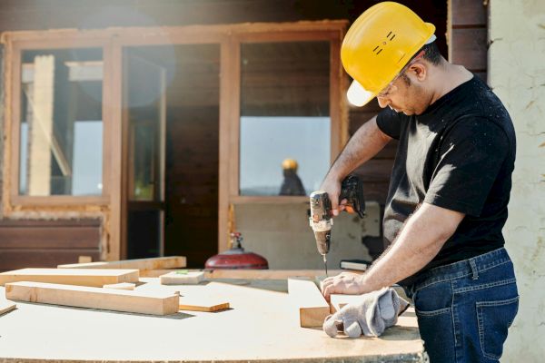 A man, wearing a yellow hard hat and black shirt, uses a power drill on wooden pieces at a construction site, with a building in the background.