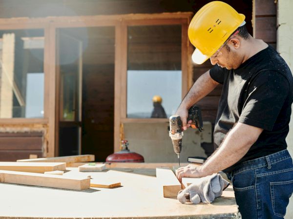 A man, wearing a yellow hard hat and black shirt, uses a power drill on wooden pieces at a construction site, with a building in the background.