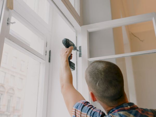 A man is using a power drill to fix or install a window frame in a room with bright natural light coming through.