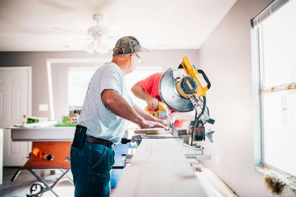 Two people are working with a miter saw on a piece of wood in a well-lit room, appearing to be engaged in a home improvement project.