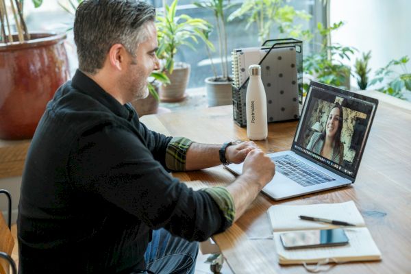 A man is sitting at a desk, engaging in a video call on his laptop. There are plants, a water bottle, and a notebook on the desk.