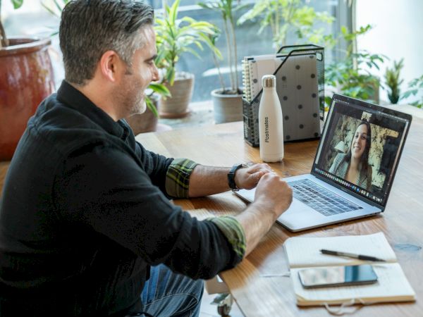 A man is sitting at a desk, engaging in a video call on his laptop. There are plants, a water bottle, and a notebook on the desk.