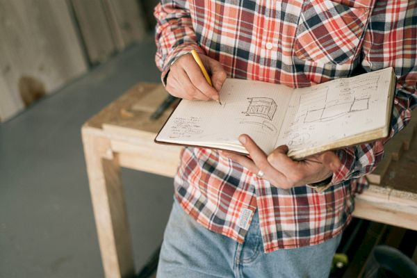 A person in a plaid shirt and jeans is sketching designs in a notebook while standing by a wooden workbench in a workshop.