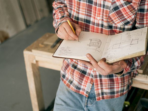 A person in a plaid shirt and jeans is sketching designs in a notebook while standing by a wooden workbench in a workshop.