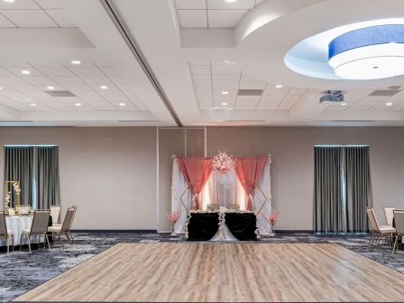 An elegant event space with a wooden dance floor, tables with chairs, and a decorated stage with pink drapes and floral arrangements.