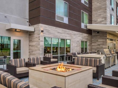 An outdoor seating area with striped cushioned chairs surrounds a modern fire pit. Nearby are grills and a modern apartment or hotel building.