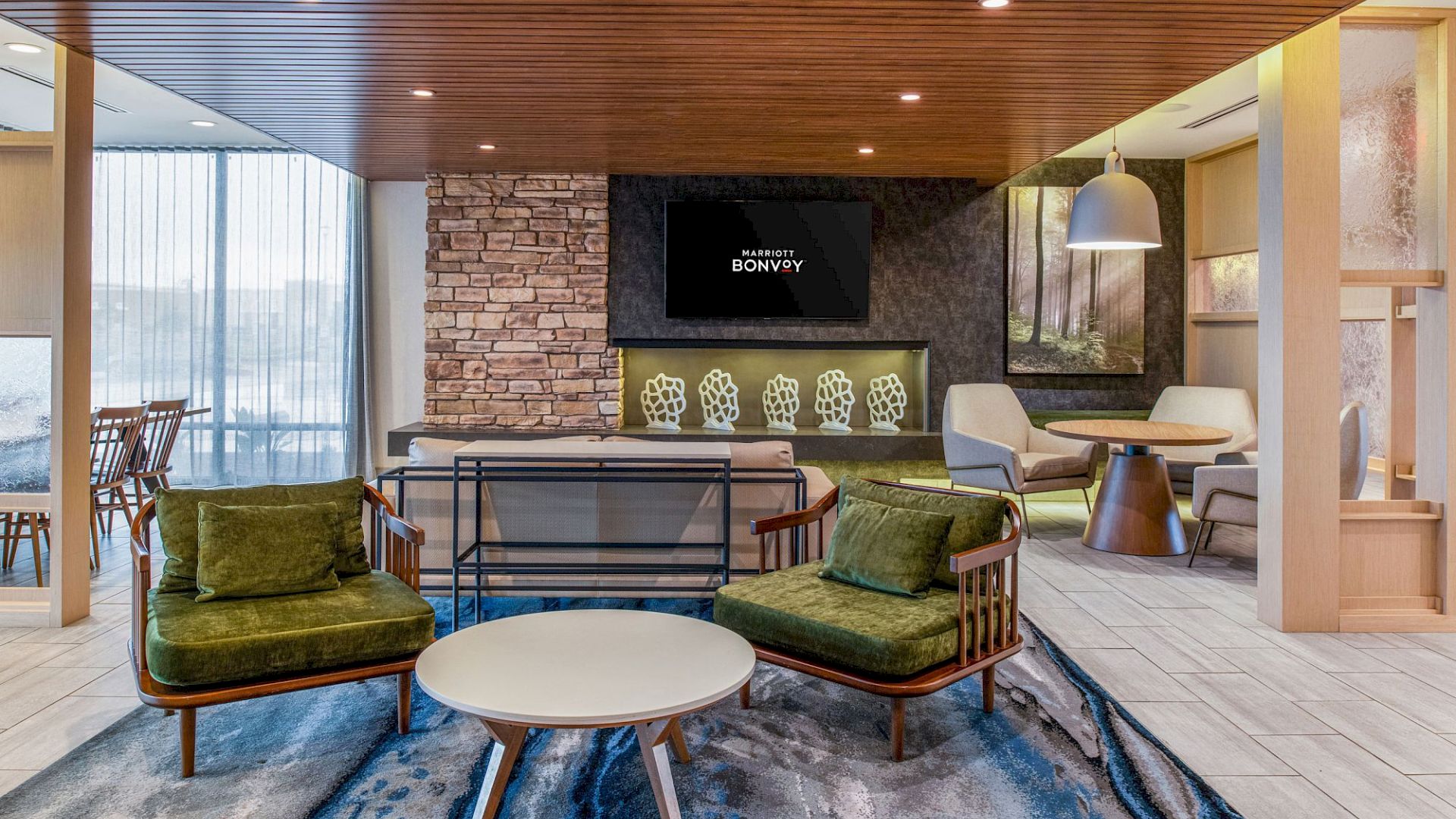 The image shows a modern living area with green chairs, a round coffee table, a television, and a fireplace, featuring stone and wood elements.