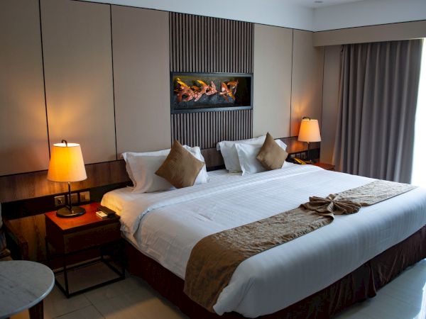 A modern hotel room with a large bed, decorative pillows, nightstands, lamps, and a wall-mounted fireplace above the headboard.