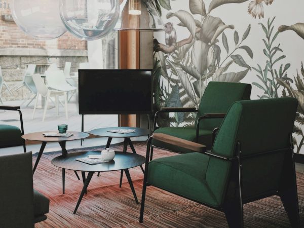 A modern living room with green chairs, a TV, a coffee table, and a botanical wallpaper, illuminated by large pendant lights.