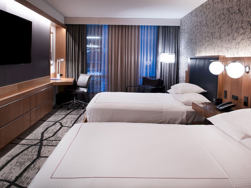 The image shows a modern hotel room with two neatly made beds, a TV, a desk with a chair, and contemporary decor including wall patterns and lighting.