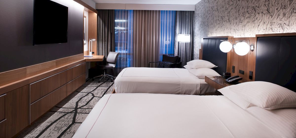 The image shows a modern hotel room with two neatly made beds, a TV, a desk with a chair, and contemporary decor including wall patterns and lighting.
