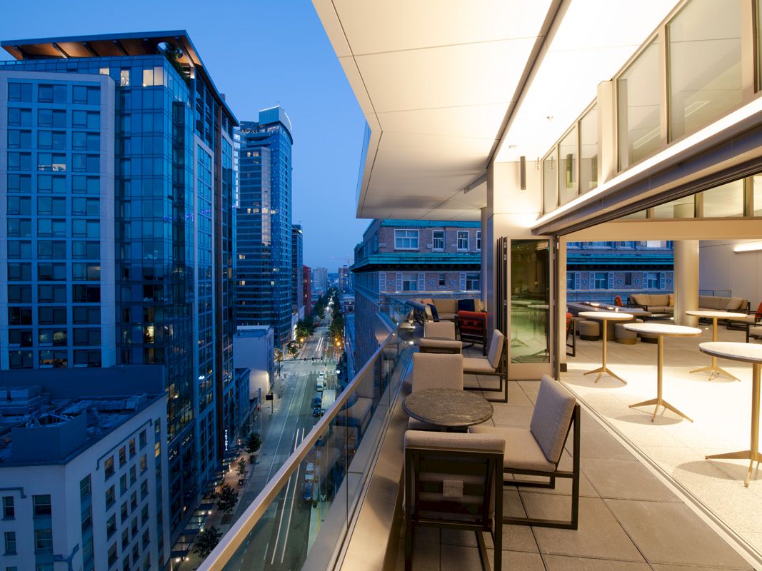 A modern balcony with tables and chairs overlooks a city street lined with tall buildings at dusk, featuring a sleek urban ambiance.