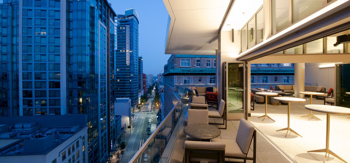 A modern balcony with tables and chairs overlooks a city street lined with tall buildings at dusk, featuring a sleek urban ambiance.