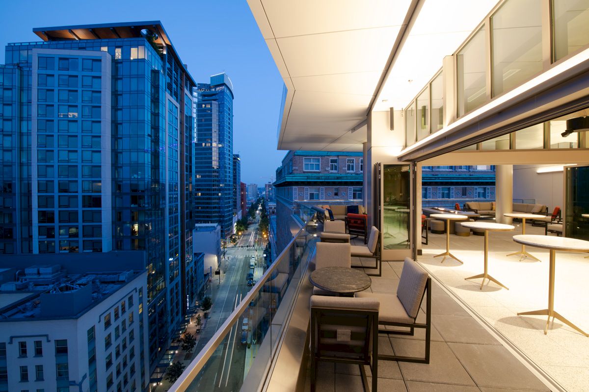 The image shows a modern cityscape viewed from a high balcony, with stylish outdoor seating and tables, overlooking tall buildings at dusk.
