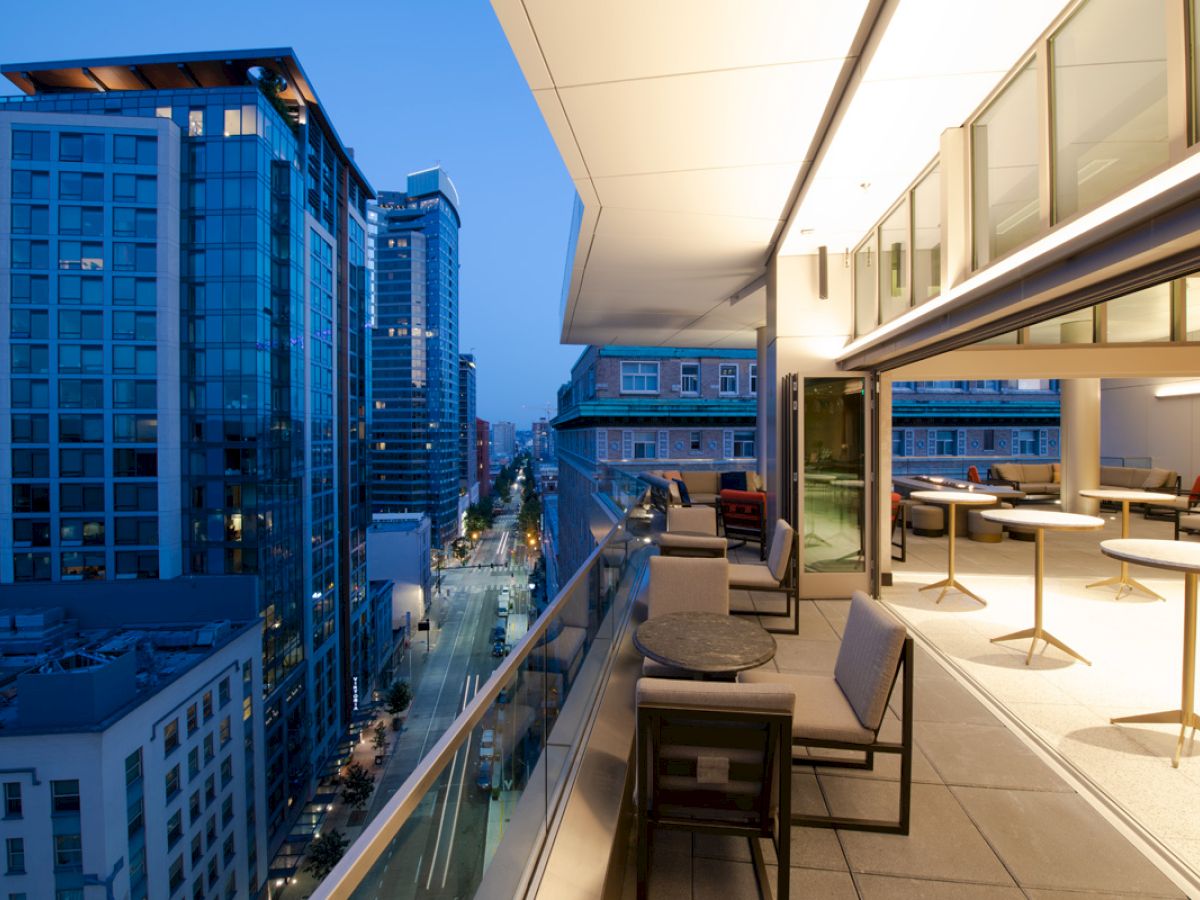 The image shows a modern cityscape viewed from a high balcony, with stylish outdoor seating and tables, overlooking tall buildings at dusk.