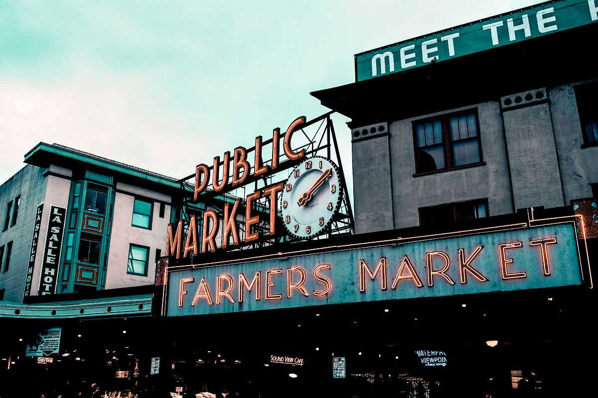 The image shows a neon sign for a "Public Market" and "Farmers Market" with a clock, in front of urban buildings, possibly a historic site.