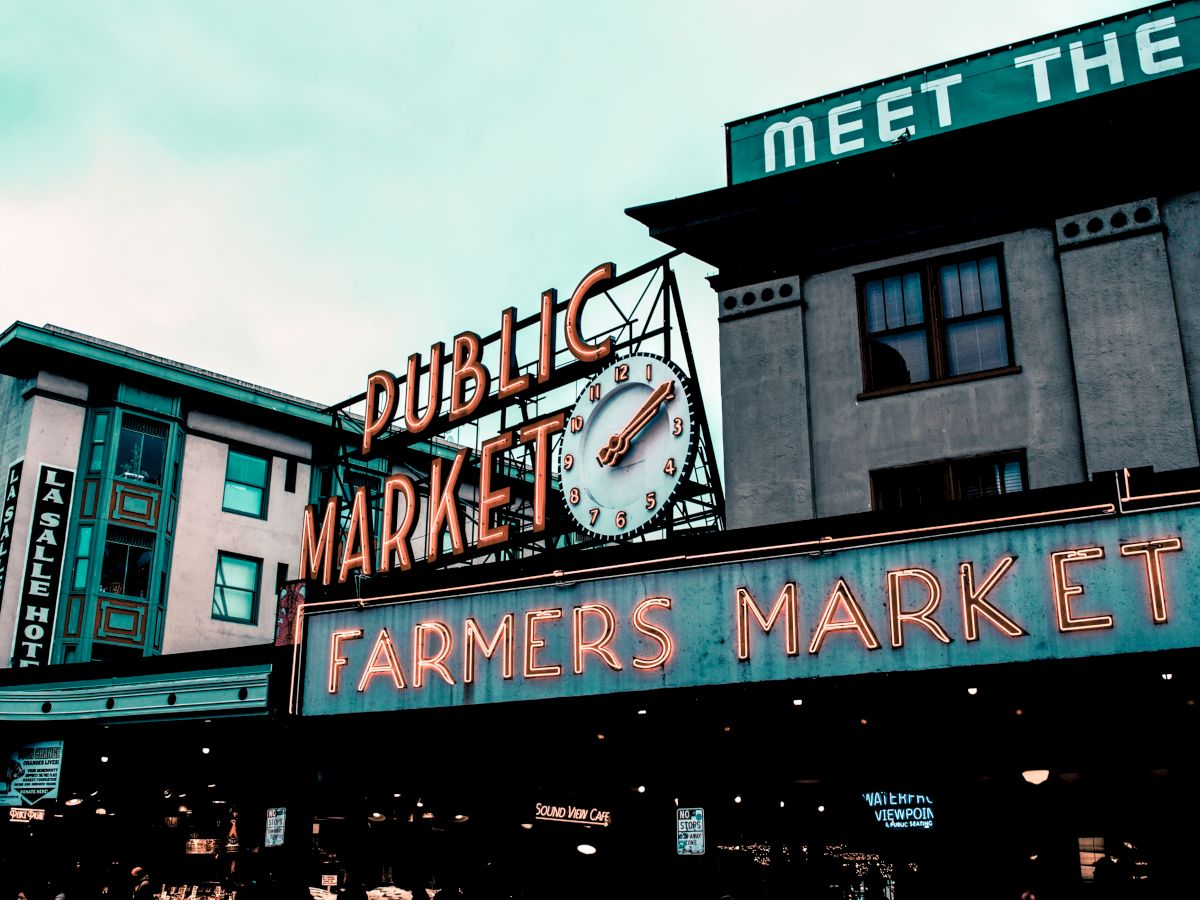 The image shows a neon sign for a "Public Market" and "Farmers Market" with a clock, in front of urban buildings, possibly a historic site.