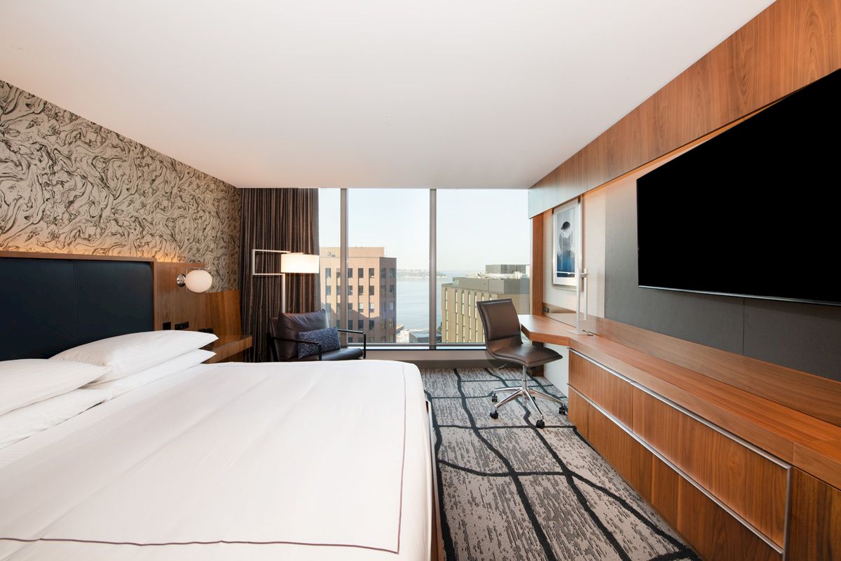 This image shows a modern hotel room with a large bed, desk, chair, wall-mounted TV, and window with a city view.
