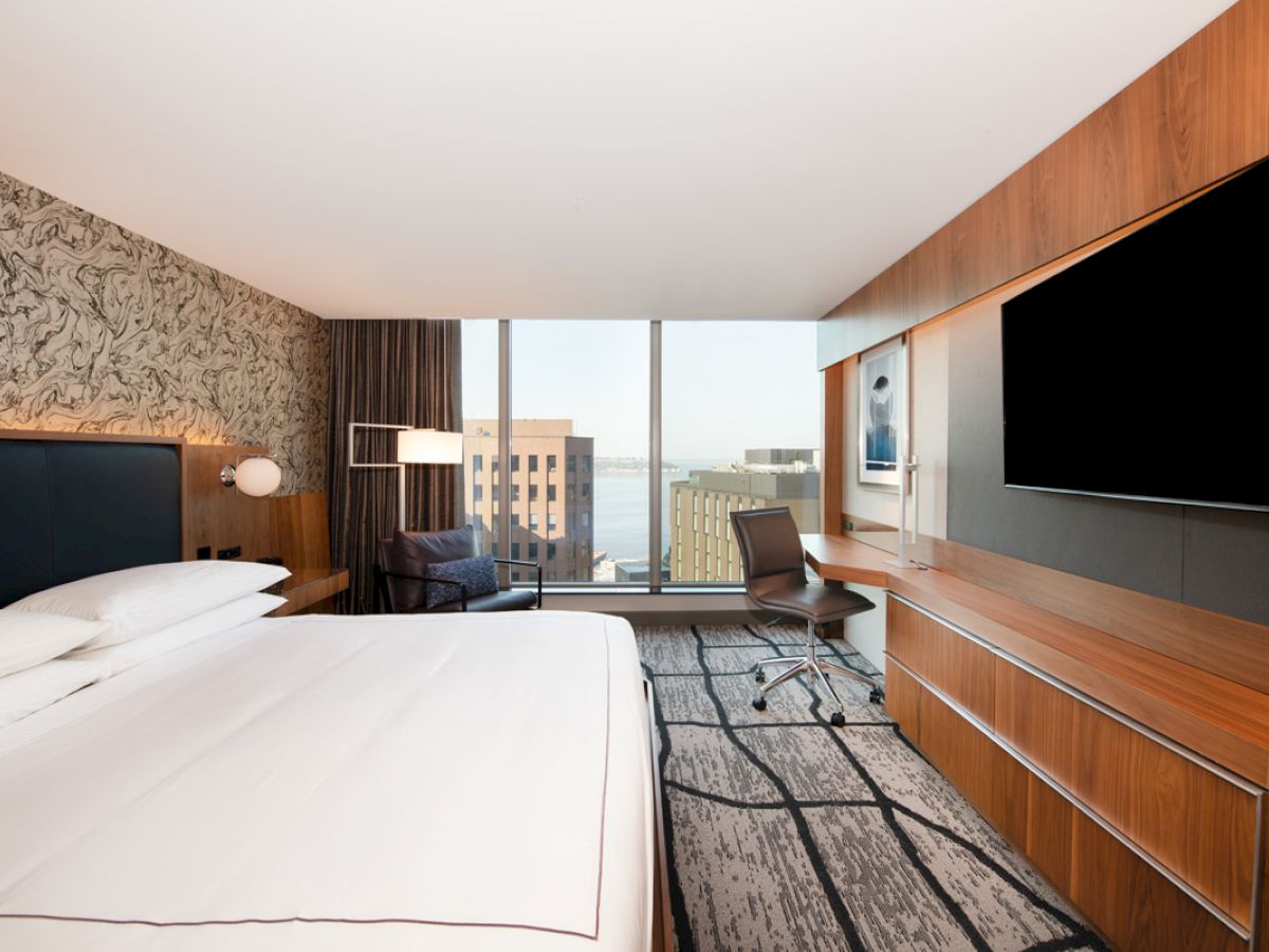 This image shows a modern hotel room with a large bed, desk, chair, wall-mounted TV, and window with a city view.