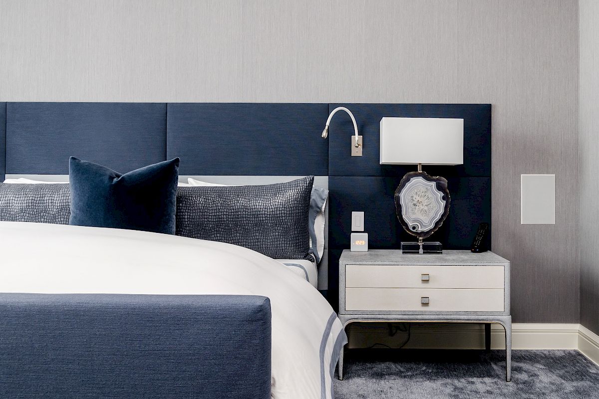 A modern bedroom with a blue upholstered bed, a nightstand with a lamp, and a decorative pillow arrangement, all set against a grey wall.