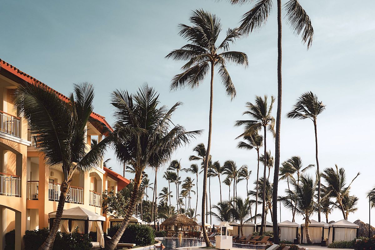 A luxurious resort pool area with palm trees, lounge chairs, and a building in the background, set under a clear sky.
