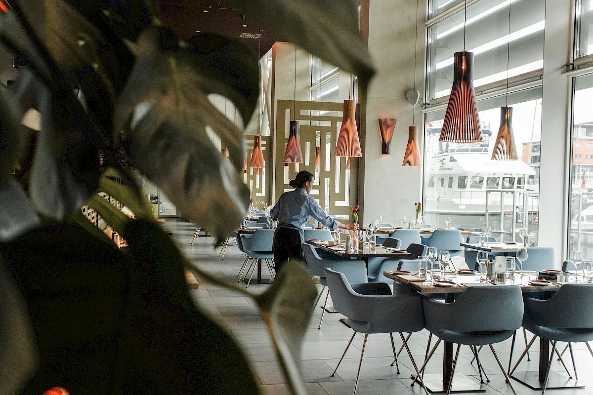 A person is setting up tables in a modern restaurant, featuring large windows and stylish lighting fixtures.