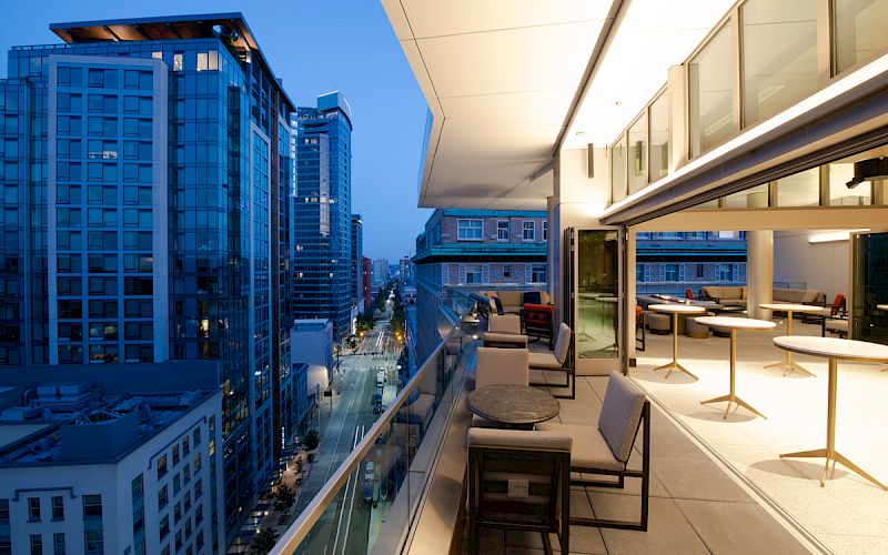 A modern outdoor balcony with seating areas overlooks a cityscape of tall buildings and streets at dusk, featuring a sleek and contemporary design.