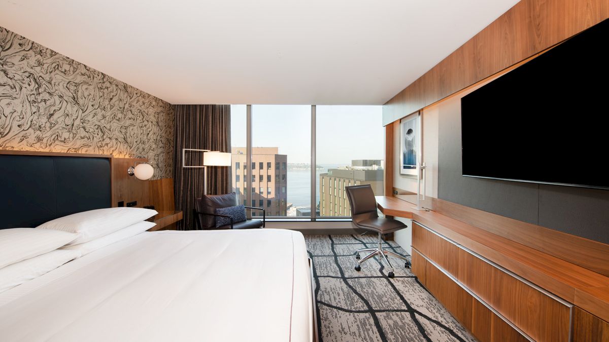A modern hotel room features a large bed, wall-mounted TV, desk with chair, and a view of city buildings through floor-to-ceiling windows.